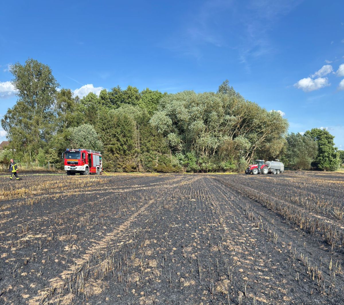 Verkohltes Stoppfelfeld mit Feuerwerhfahrzeug und Traktor mit Wassersilo im Hintergrund.