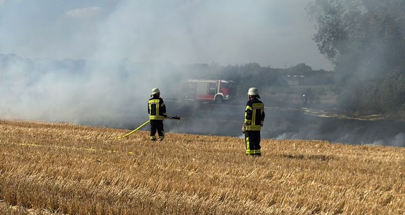 2 Feuerwehrleute auf dem ausgebrannten Feld mit Rauchwand. Im Hintergrund ist ein Feuerwehrfahrzeug.