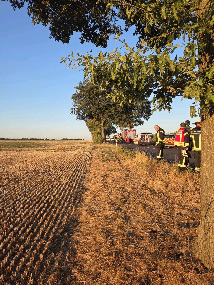 Blick auf ein trockenes Stoppelfeld mit Feuerwehrleuten und Feuerwehrautos am Straßenrand.